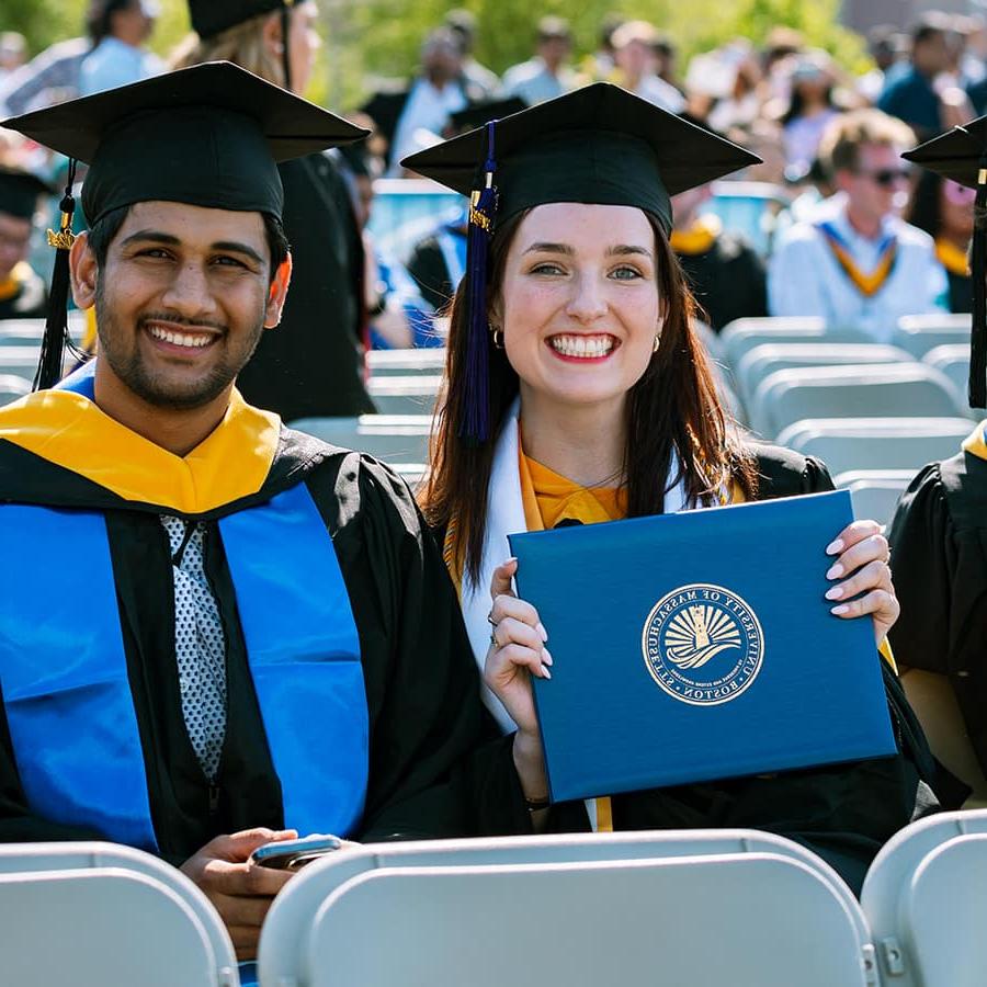 2 sit outside at commencement in caps & gowns, 1 holds up diploma.